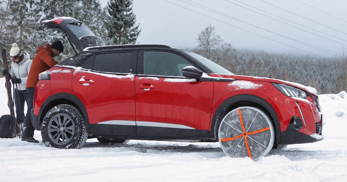 A red Peugeot SUV parked in deep snow with its trunk open, showing two people standing behind the vehicle. The car has black wheels with orange snow socks for tires installed, and the background features a snowy forest under an overcast sky.