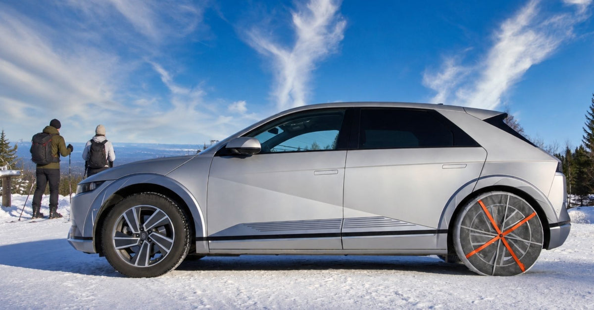 A silver electric crossover SUV with snow chains mounted on rear wheels and parked in a snowy outdoor setting under a bright blue sky with wispy clouds. Two people are standing in the snow in the background near trees.