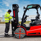 A red and black Manitou forklift parked outdoors on asphalt, with a worker in a high-visibility yellow jacket standing beside the mast and forks under a blue sky. Snow socks are mounted on front wheels.
