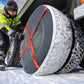 A truck driver in a high-visibility winter jacket is attaching a bright orange strap snow sock to a large winter tire on a truck tire in the snow.