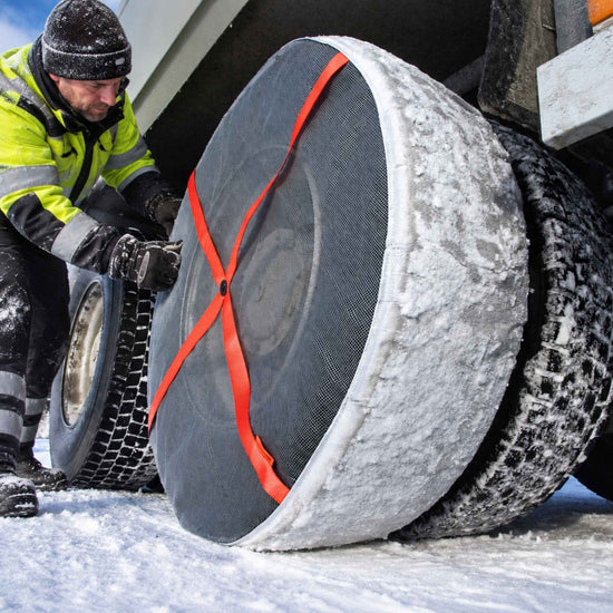 A truck driver in a high-visibility winter jacket is attaching a bright orange strap snow sock to a large winter tire on a truck tire in the snow.