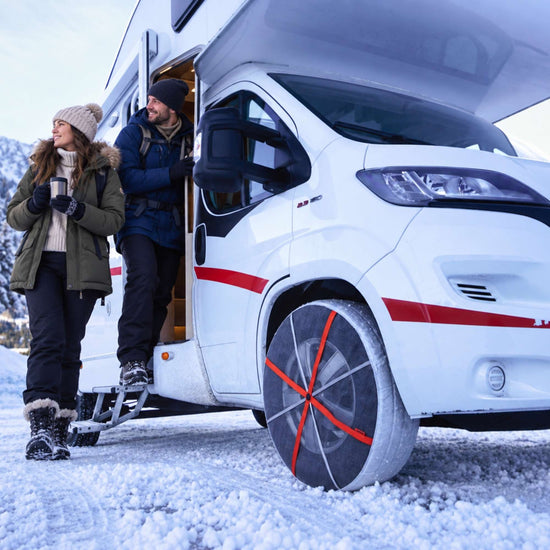 A white motorhome parked in the snow with two people standing by the open side door. The front tire is fitted with snow socks that have orange crossed straps.