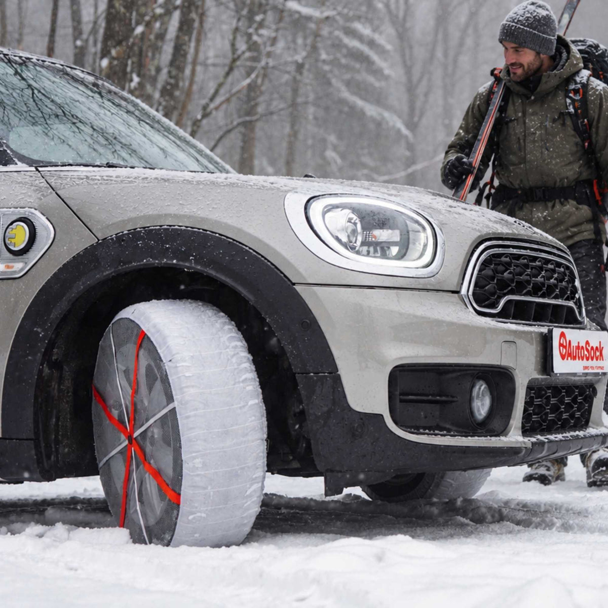 A silver Mini Cooper in a snowy outdoor setting, showing a winter tire chain installed on the front wheel. A man in winter gear and ski boots stands in the background near the car.