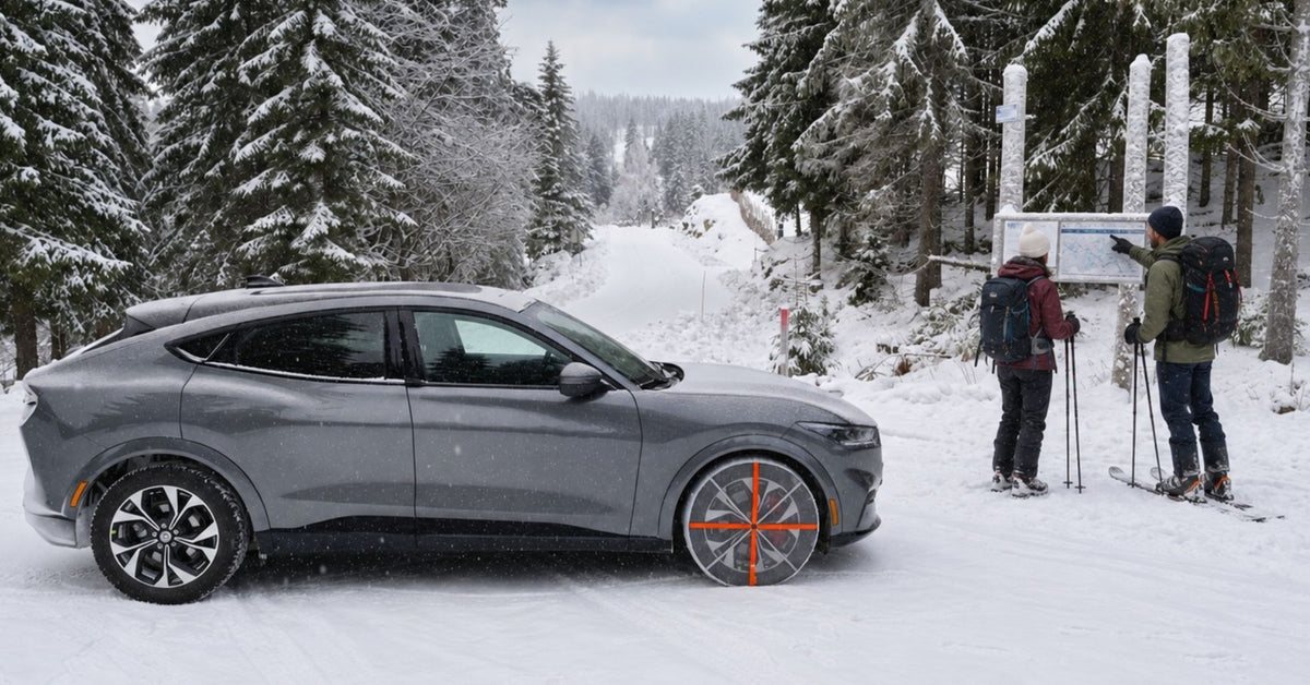 A gray Ford Mustang SUV with textile snow chains mounted and parked on a snowy mountain road with skiers standing nearby under snow-covered pine trees.