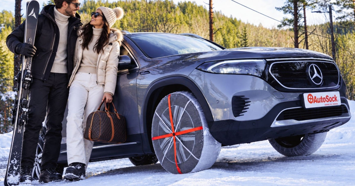A gray Mercedes-Benz electric SUV with snow socks installed on winter tires, parked in a snowy outdoor setting with two people standing beside it, one holding skis and the other holding a brown handbag. 